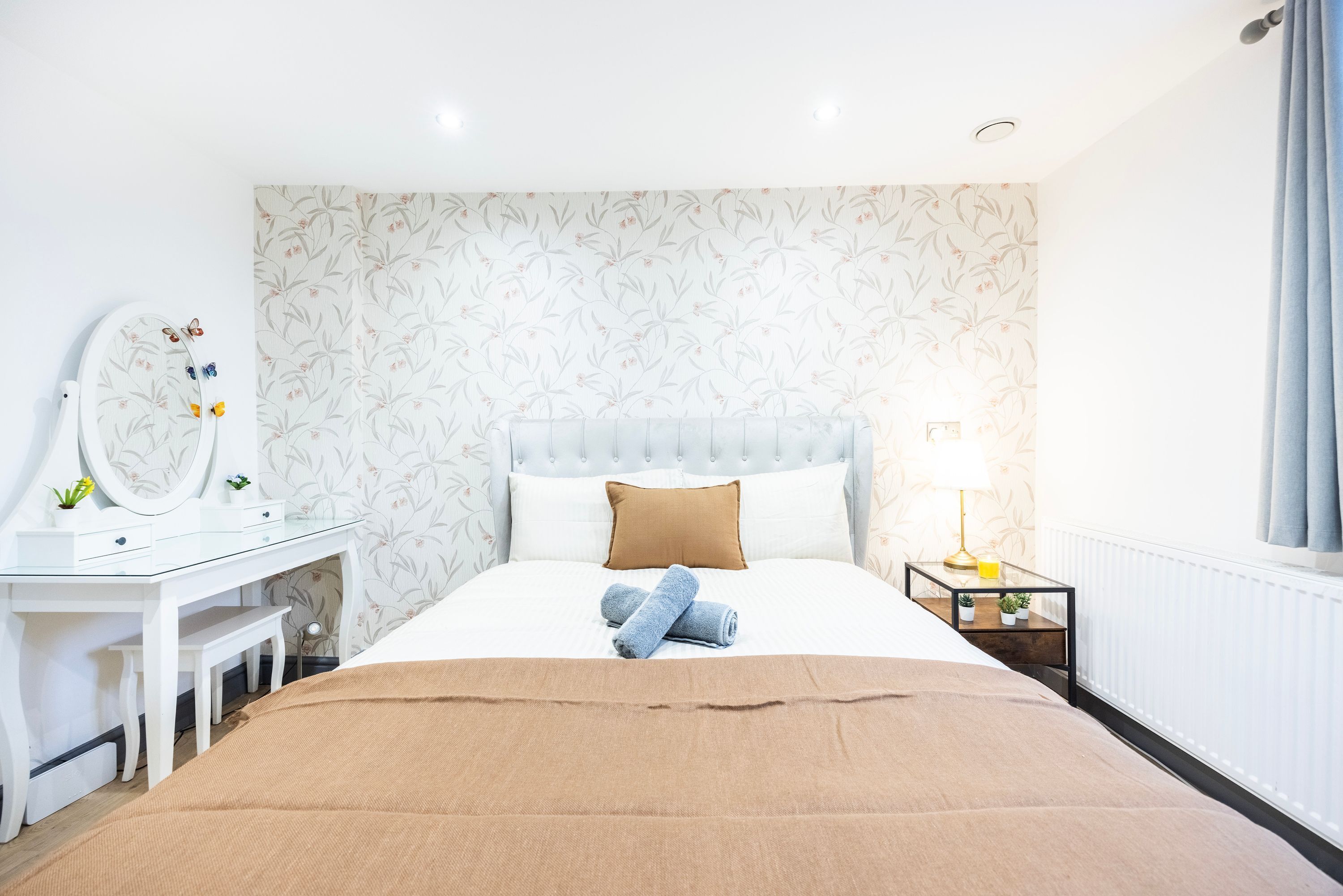 A neatly arranged bedroom with a bed, a bedside table with a lamp, a dresser with a mirror, and a wall with a floral pattern.