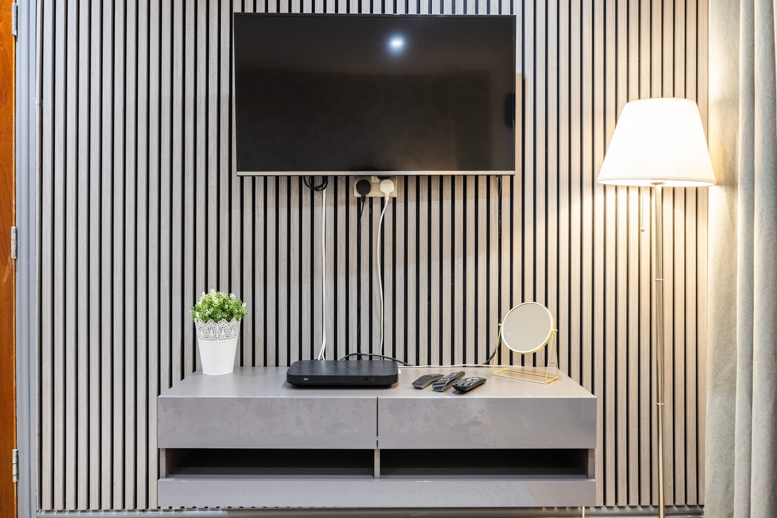 A modern living room with a flat-screen TV mounted on a wooden wall, a grey console table with a plant, remote controls, and a lamp.