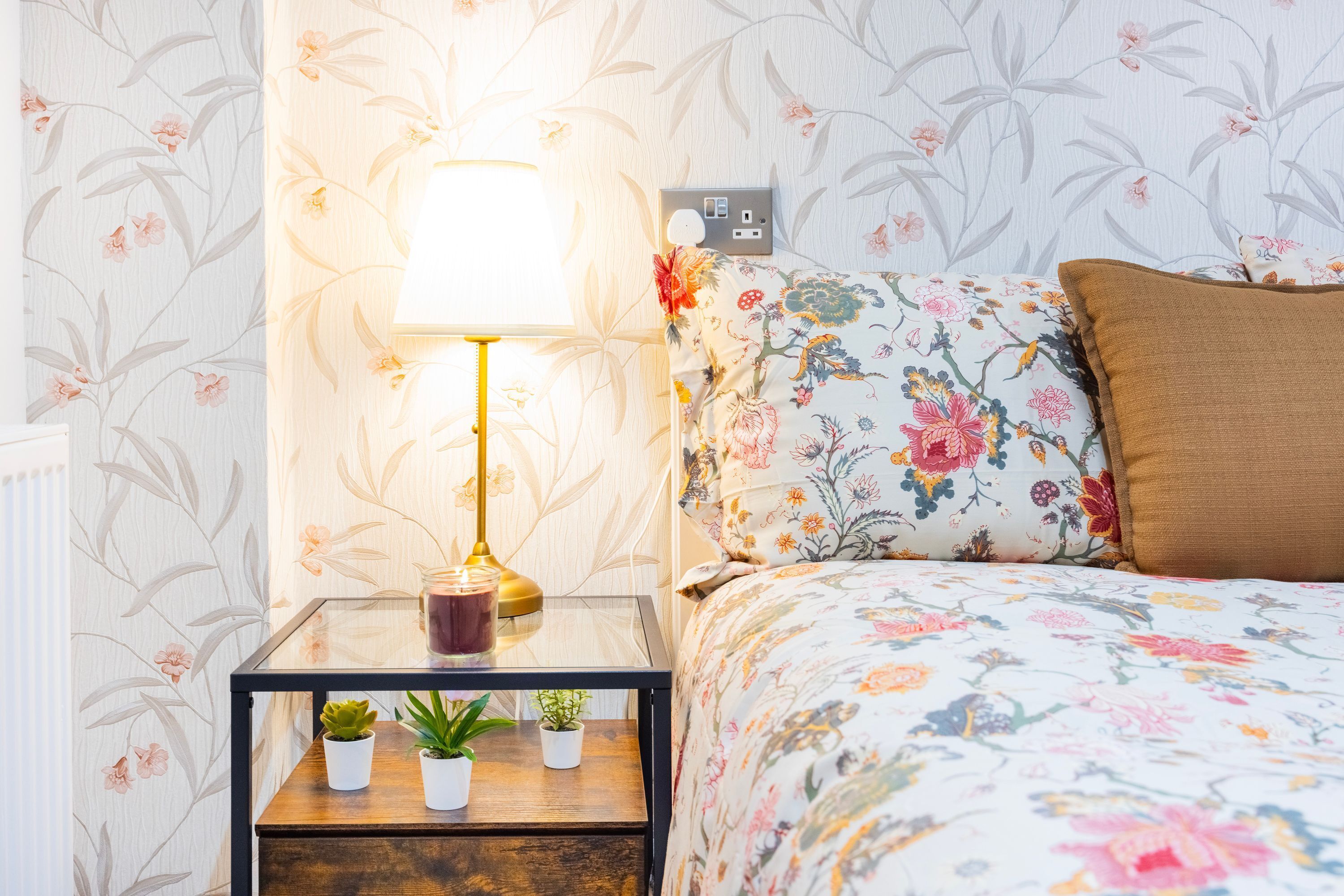 Un dormitorio con una colcha de estampado floral, una mesita de noche con lámpara y dos macetas.