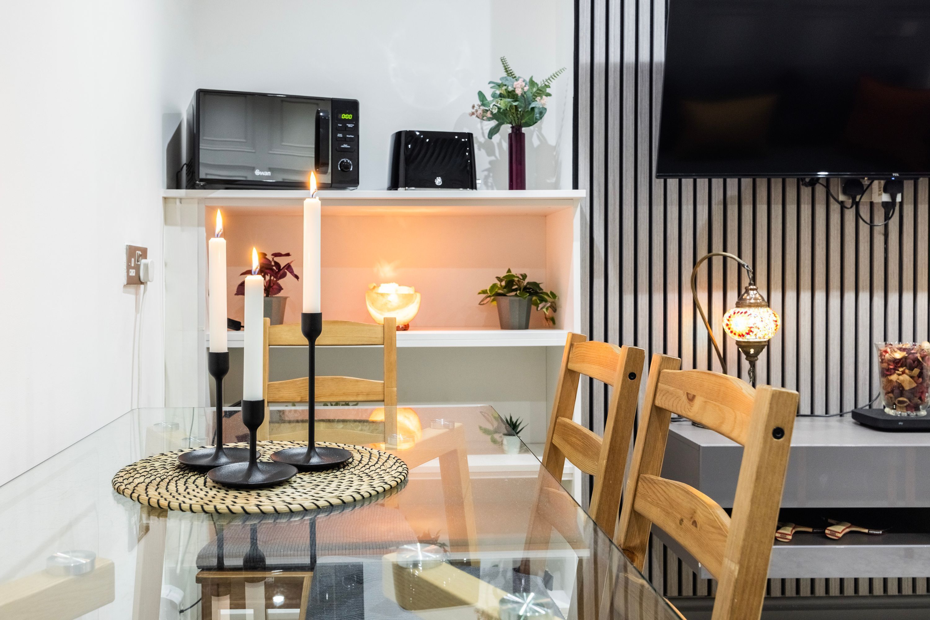 Un salon moderne avec une table à manger en verre, des chaises en bois, un téléviseur et une étagère ornée d’objets décoratifs.