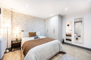 A modern bedroom with a large bed, a nightstand with a lamp, a wardrobe, and a mirror. - 3 Bedroom Apartment by River Thames (London)