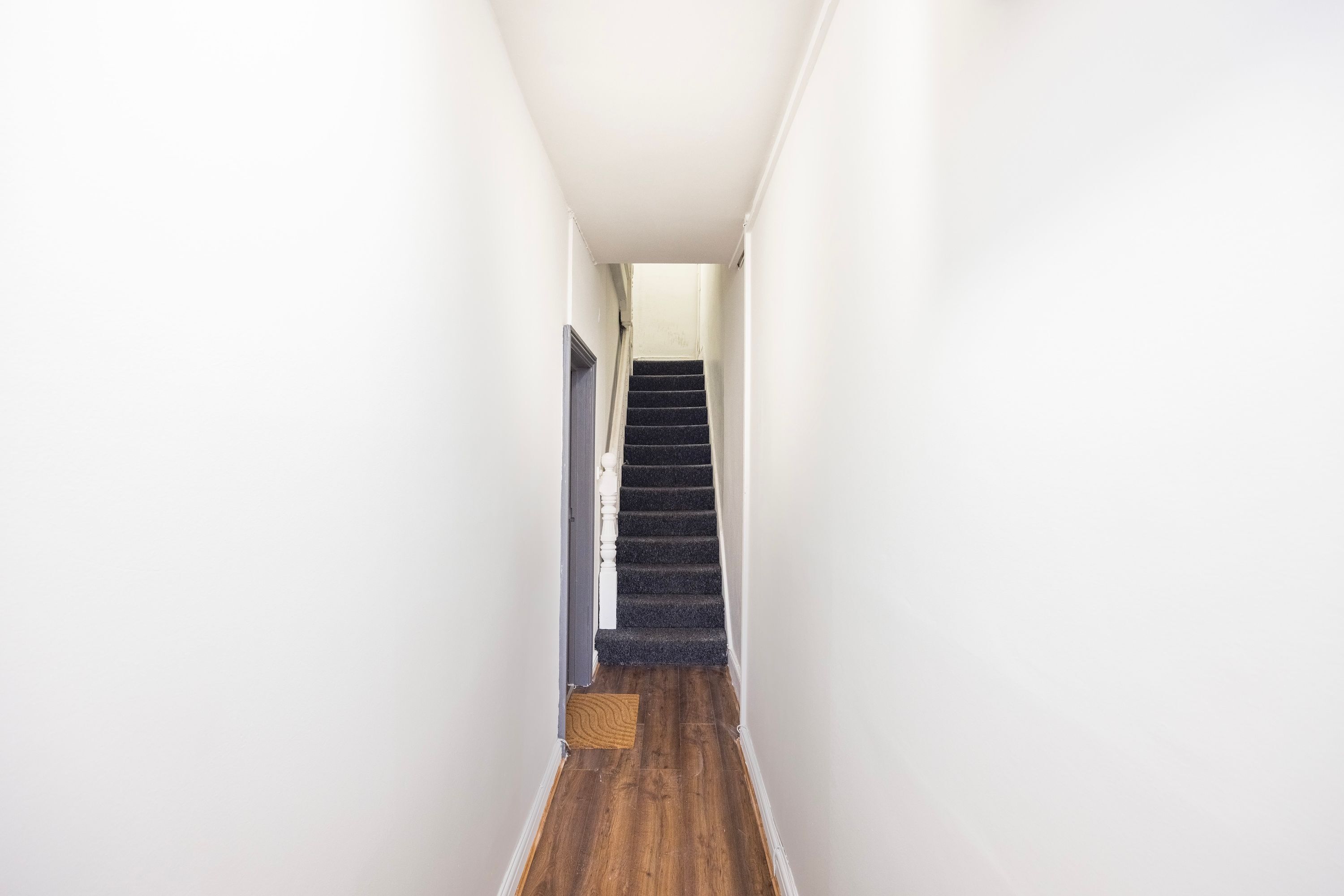 Un couloir étroit, doté d’un plancher en bois et d’un escalier recouvert de moquette, qui mène à une porte.
