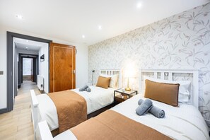 A hotel room with two beds, a wooden door, a nightstand with a lamp, and a floral wallpaper. - 3 Bedroom Apartment by River Thames (London)