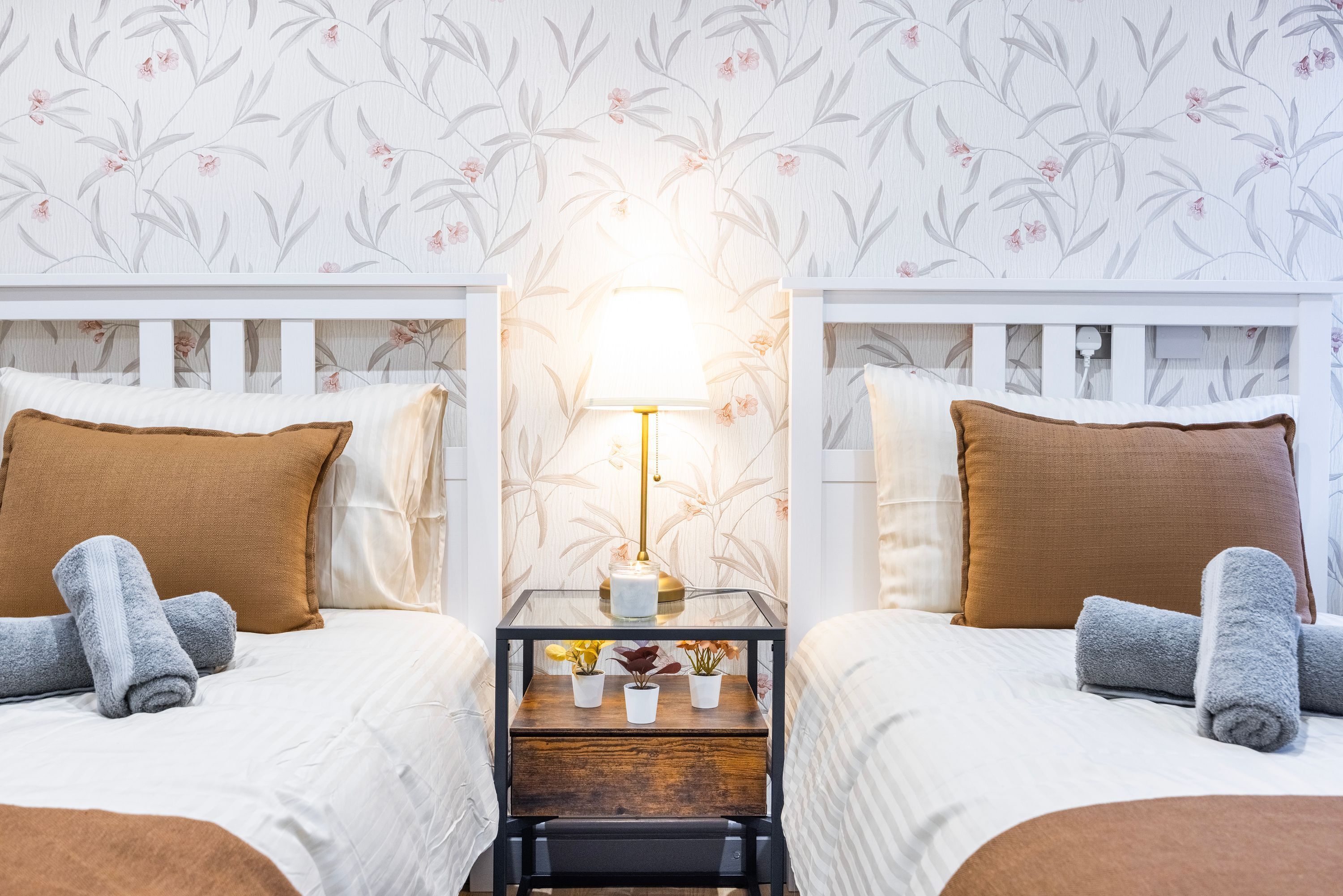 A hotel room with two beds, each with a brown pillow and a rolled gray towel. A small table with a lamp and potted plants is between the beds.