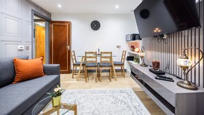 Ein modernes Wohnzimmer mit einem grauen Sofa, einem Couchtisch, einem Esstisch mit Stühlen, einem an der Wand montierten Fernseher und einer Wanduhr.