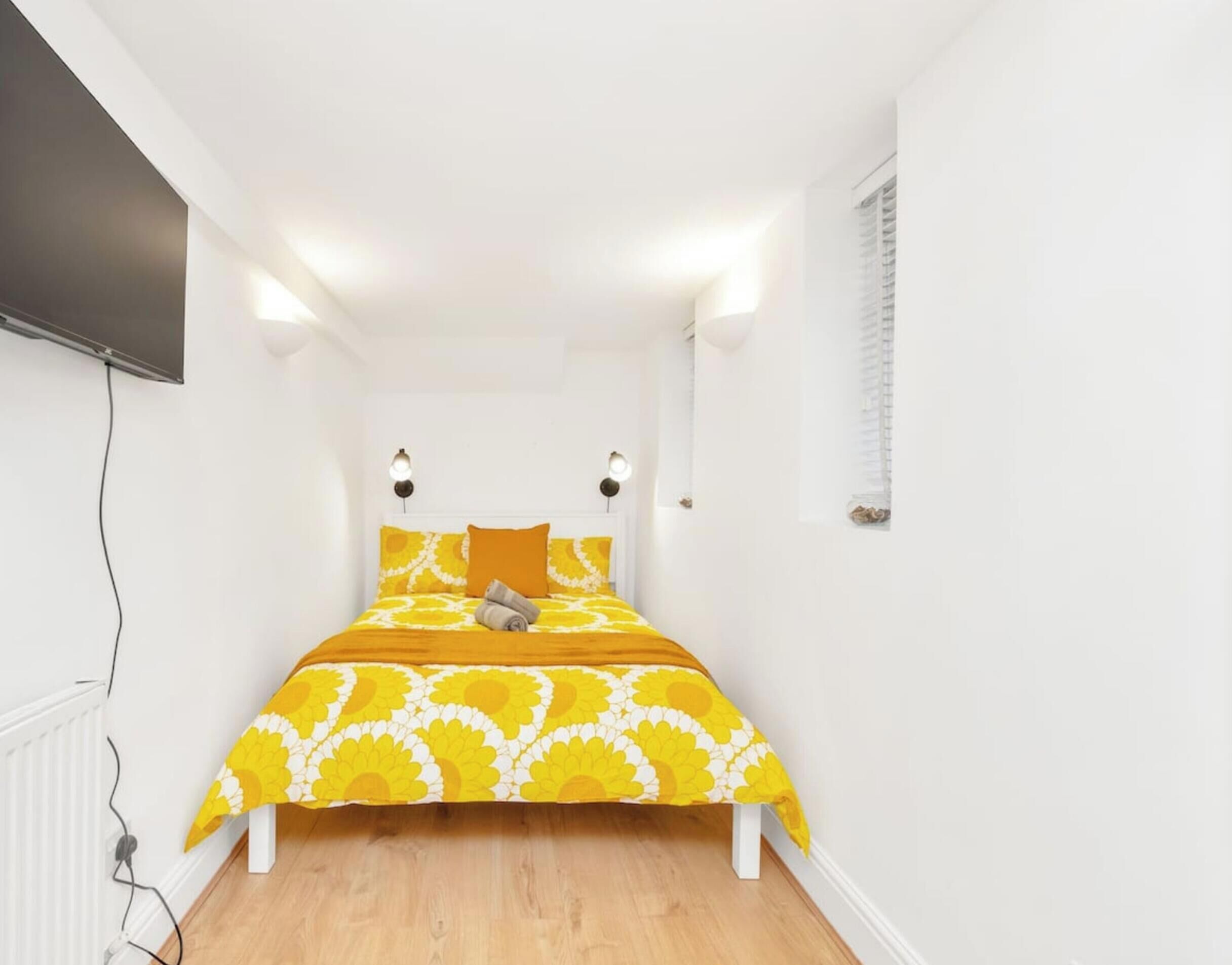 Une petite chambre lumineuse, avec un lit placé au centre, des murs blancs et une fenêtre sur le côté droit.