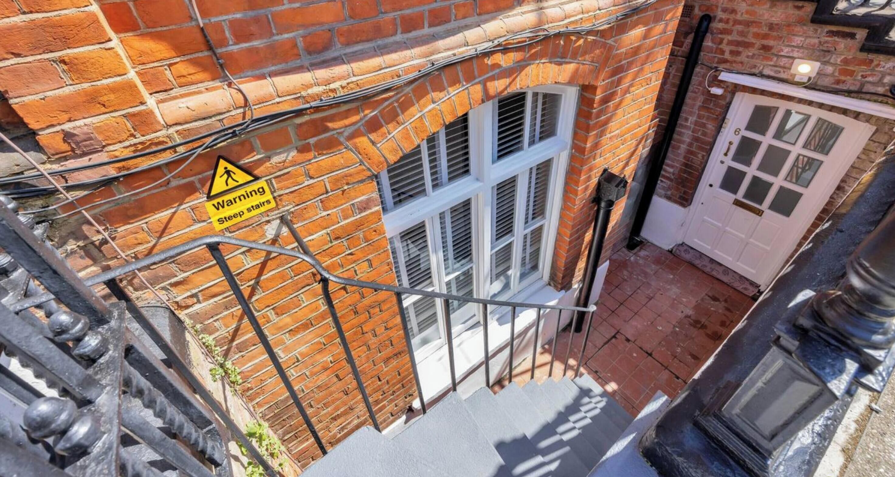 Un bâtiment en briques avec un panneau d’avertissement indiquant la présence d’un escalier raide, une porte blanche et une fenêtre avec des volets.