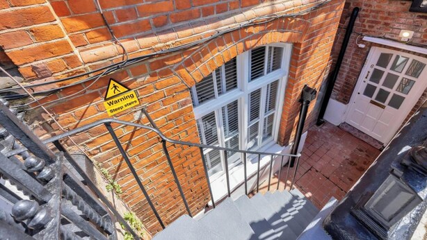Un edificio de ladrillo con un cartel de advertencia por escaleras empinadas, una puerta blanca y una ventana con contraventanas.