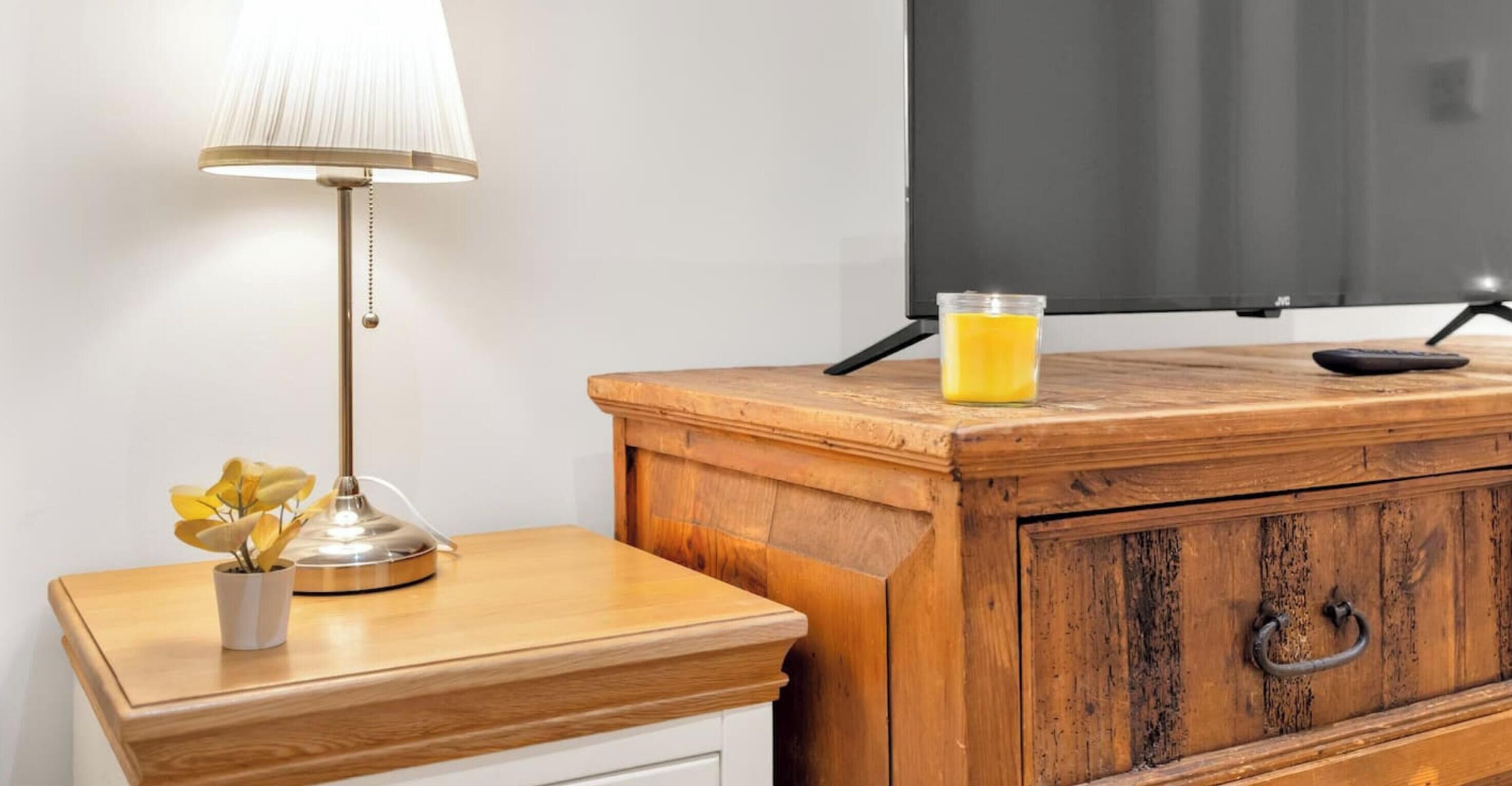 A wooden nightstand with a lamp, a small vase with flowers, a glass of juice, and a television on a stand.