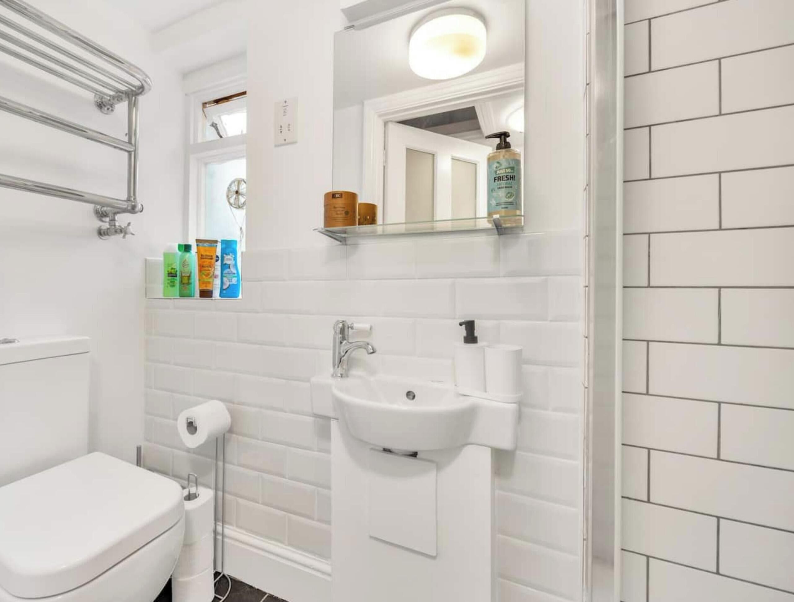 Ein Badezimmer mit Toilette, Waschbecken und Spiegel. Es befinden sich Pflegeprodukte an der Wand und ein Regal über dem Spiegel.