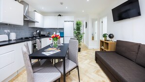 A modern kitchen with white cabinets, a black countertop, and a dining table with chairs. There is a flat-screen TV mounted on the wall and a potted plant in the corner. - 2 Bedroom Charming Family Apartment (London)