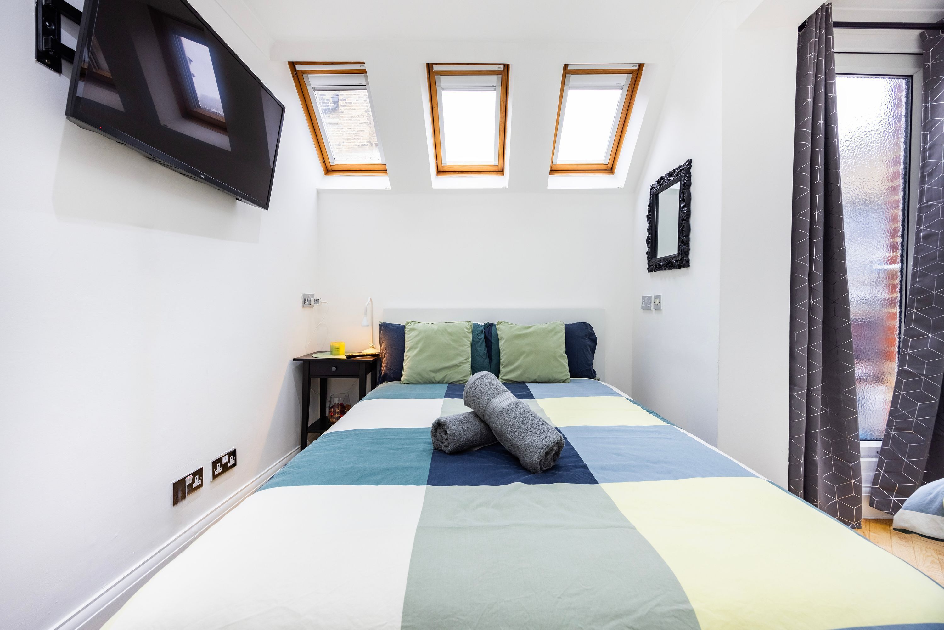 Quarto com cama, televisão de tela plana na parede, três claraboias, mesa de cabeceira com abajur e espelho na parede.