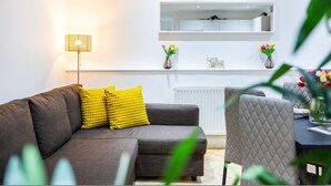 A modern living room with a grey sofa, yellow cushions, a floor lamp, and a white shelf with decorative items. - 2 Bedroom Charming Family Apartment (London)