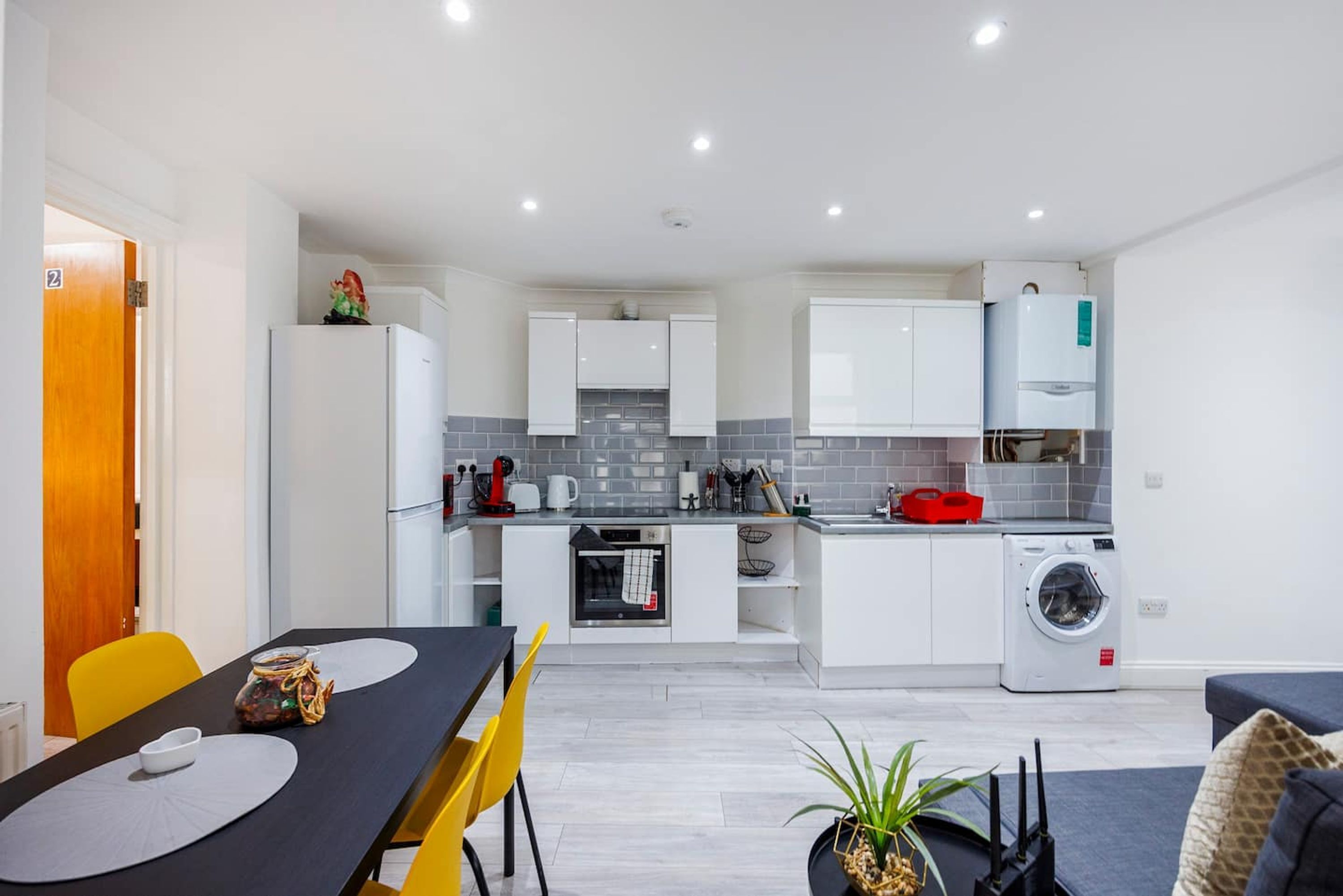 A modern kitchen with white cabinets, a built-in oven, a washing machine, and a dining area with a black table and yellow chairs.