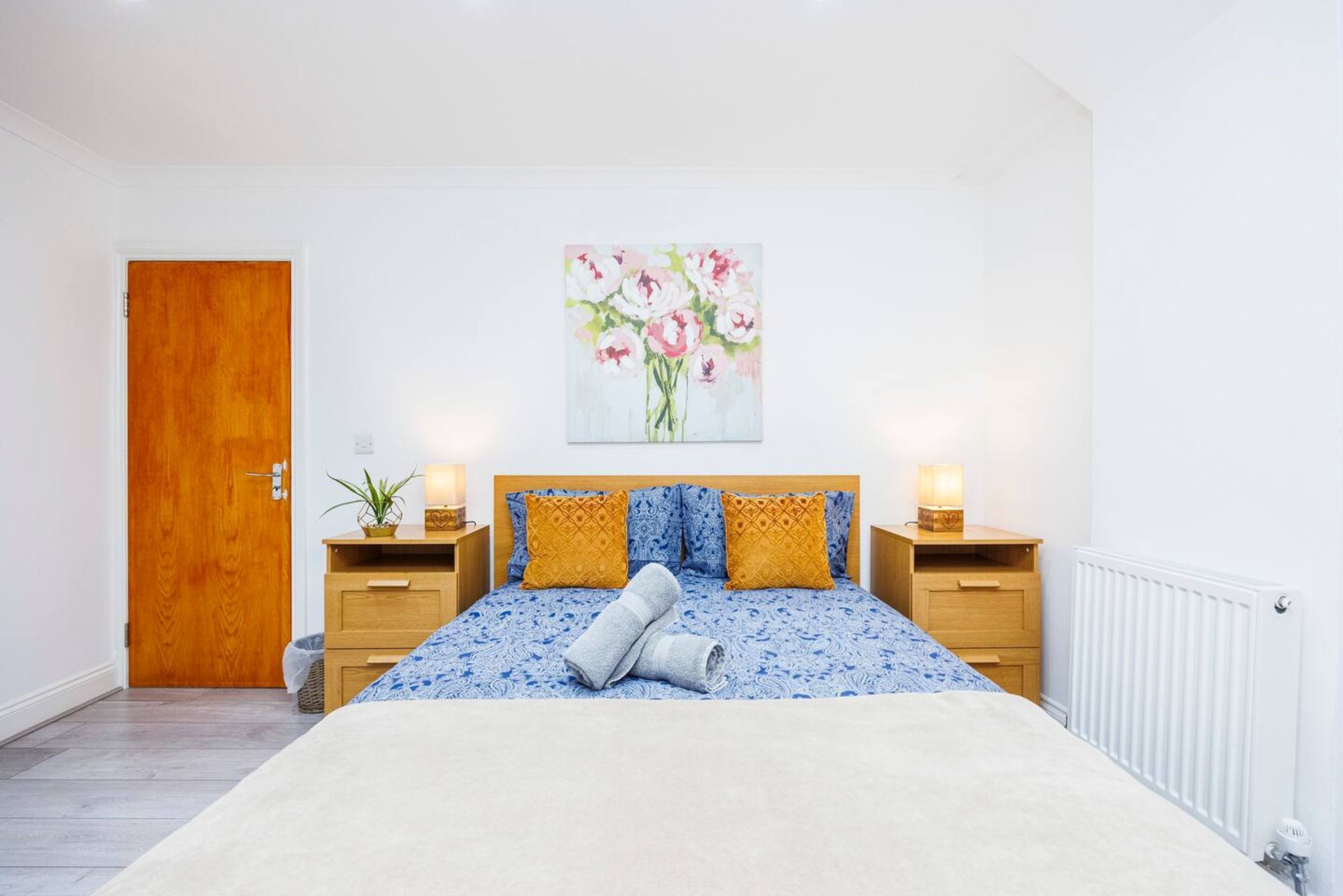 A bedroom with a wooden door, a bed with blue and yellow bedding, two bedside tables with lamps, and a painting of flowers on the wall.