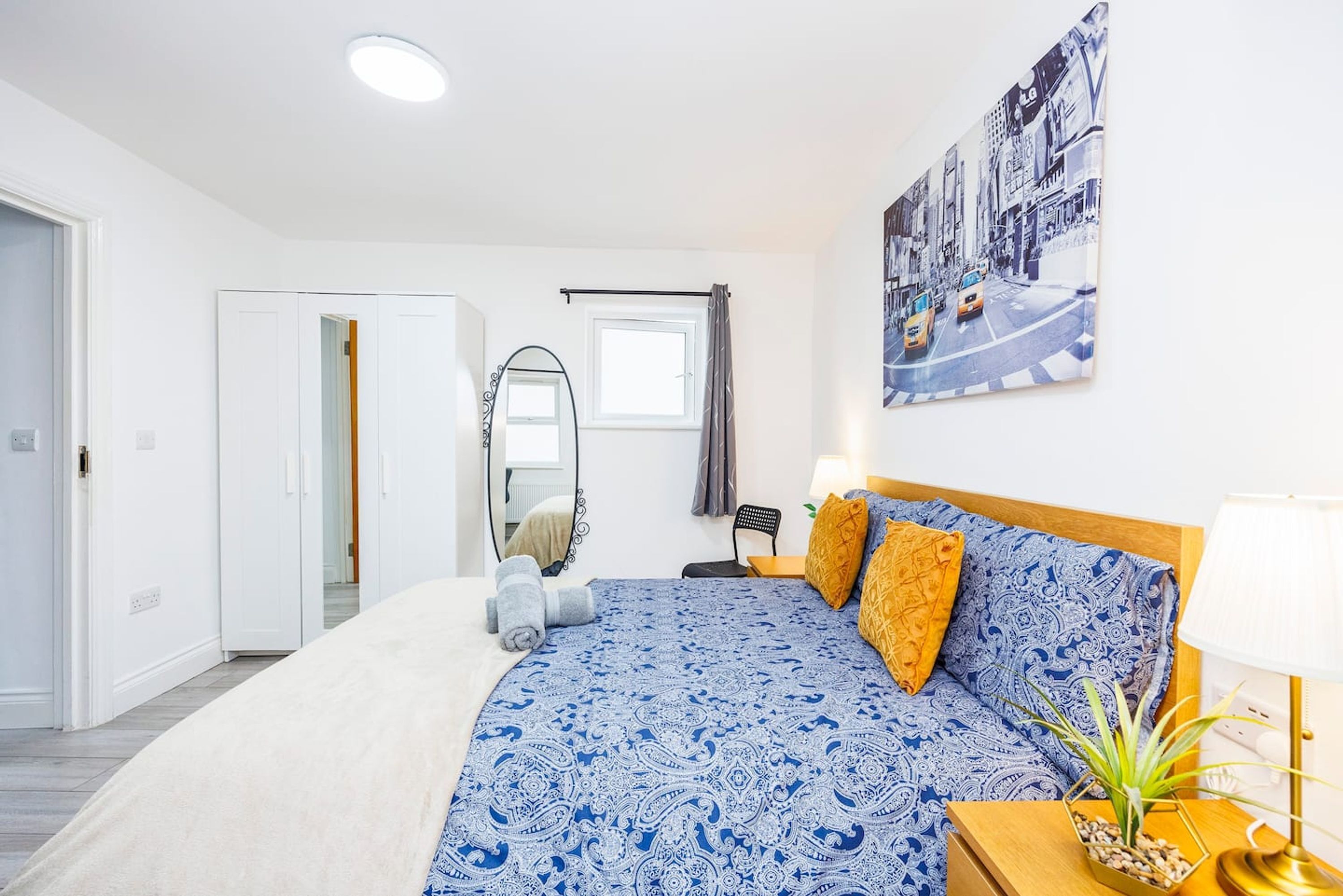 A bedroom with a bed, a bedside table with a lamp, a mirror, a window, and a cityscape painting on the wall.