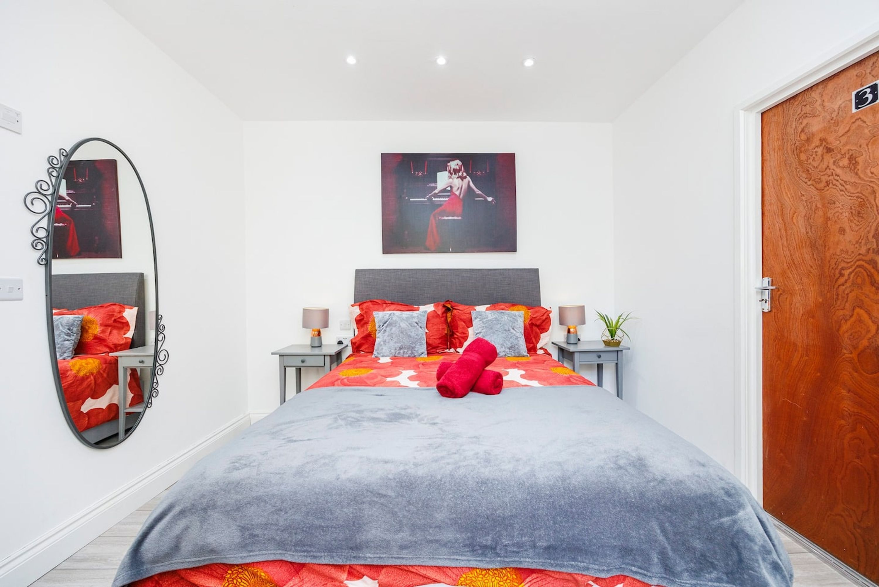 A modern bedroom with a large bed, a mirror, a framed picture on the wall, and a wooden door.