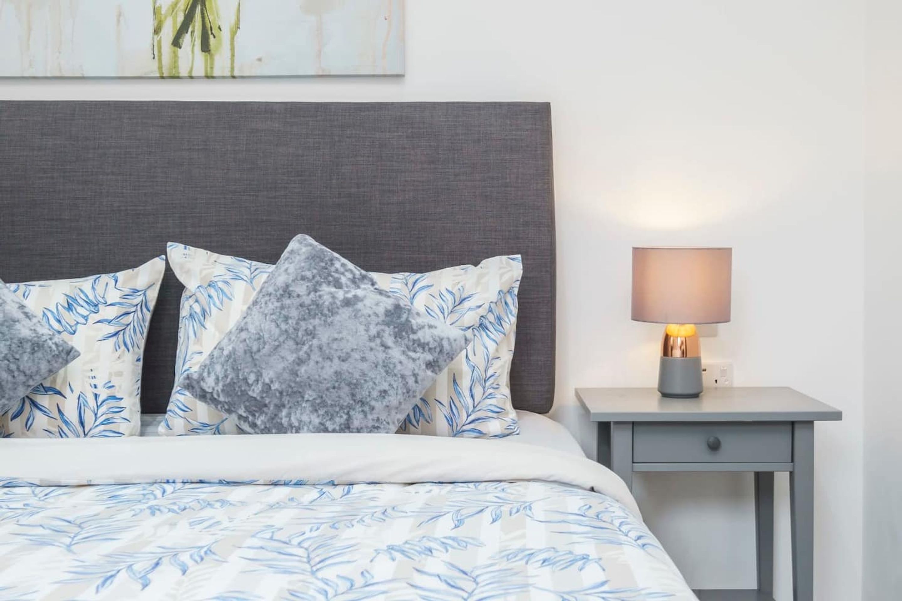 A neatly made bed with patterned bedding, a grey headboard, and a bedside table with a lamp.