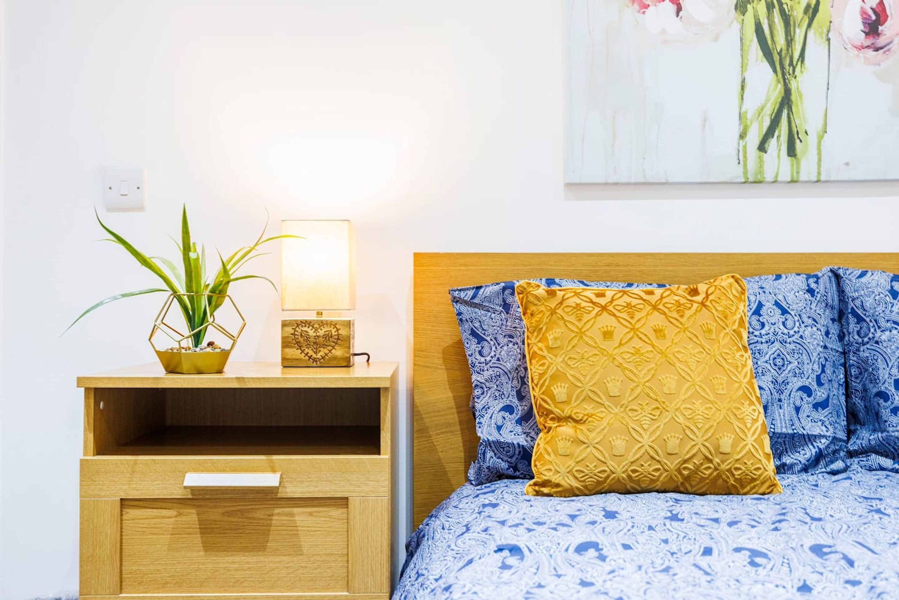 Una camera da letto ordinata con un comodino in legno, un tavolino da notte con una lampada, una pianta in vaso e un letto con lenzuola a fantasia.