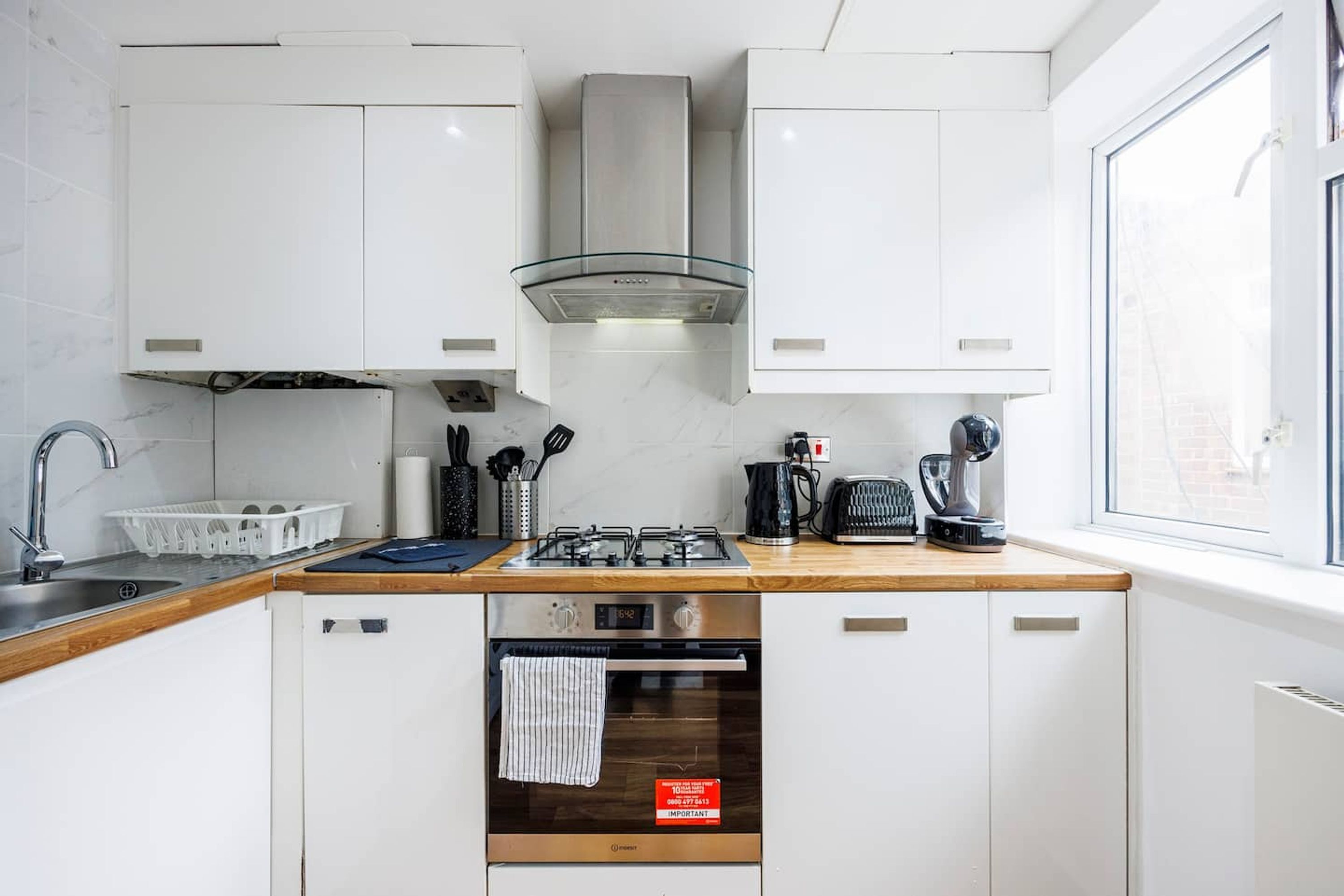 Cocina compacta con armarios blancos, extractor de acero inoxidable, horno integrado y ventana que aporta luz natural.