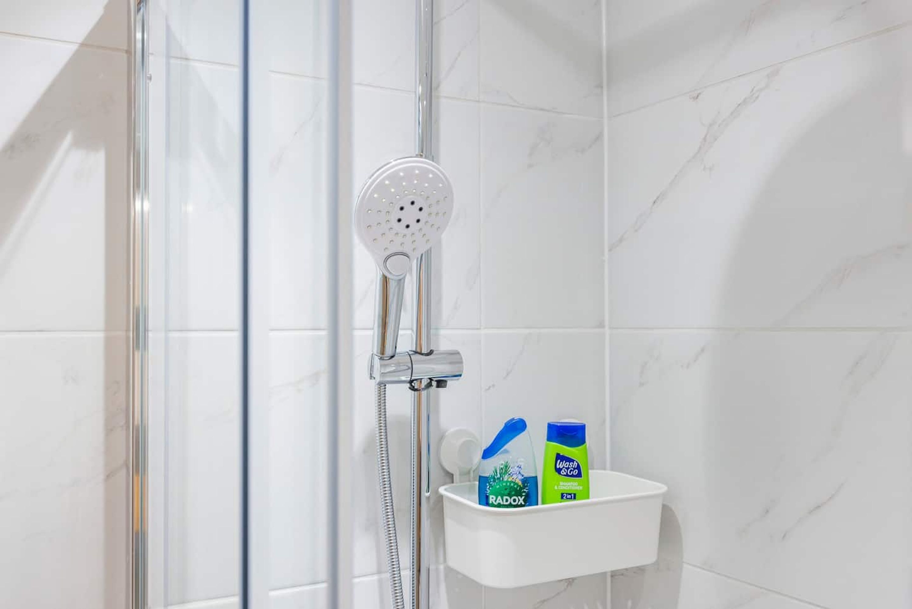 Zona de ducha con cabezal de ducha manual, puerta de cristal y estante blanco con dos botellas de Radox y un recipiente azul.