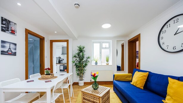 A modern living room with a blue sofa, white chairs, a wooden coffee table, and a large wall clock. - 2 Bedroom Charming Magnificent Flat (London)