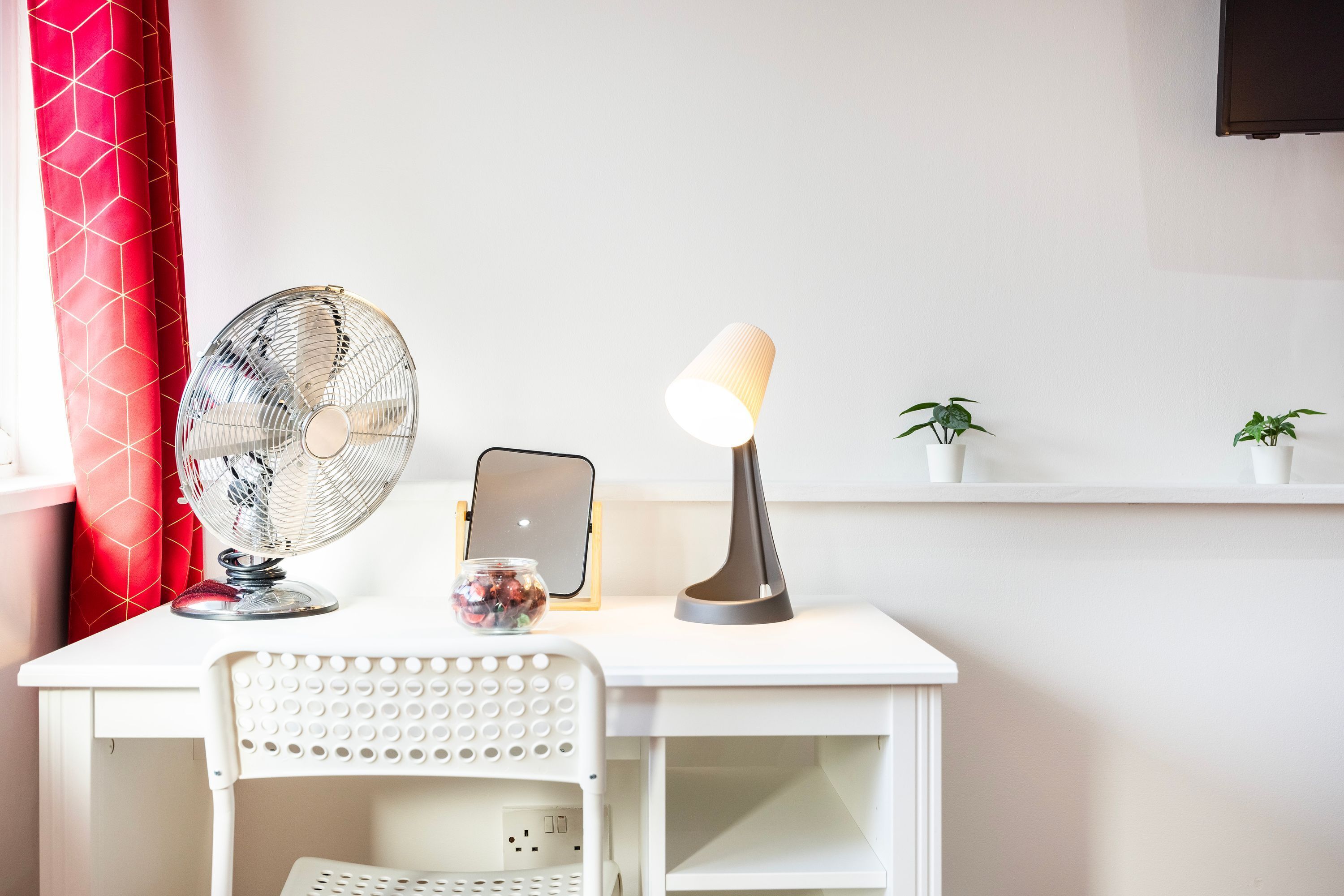 Un escritorio blanco con un ventilador de pie, una lámpara, una tableta y una planta pequeña.
