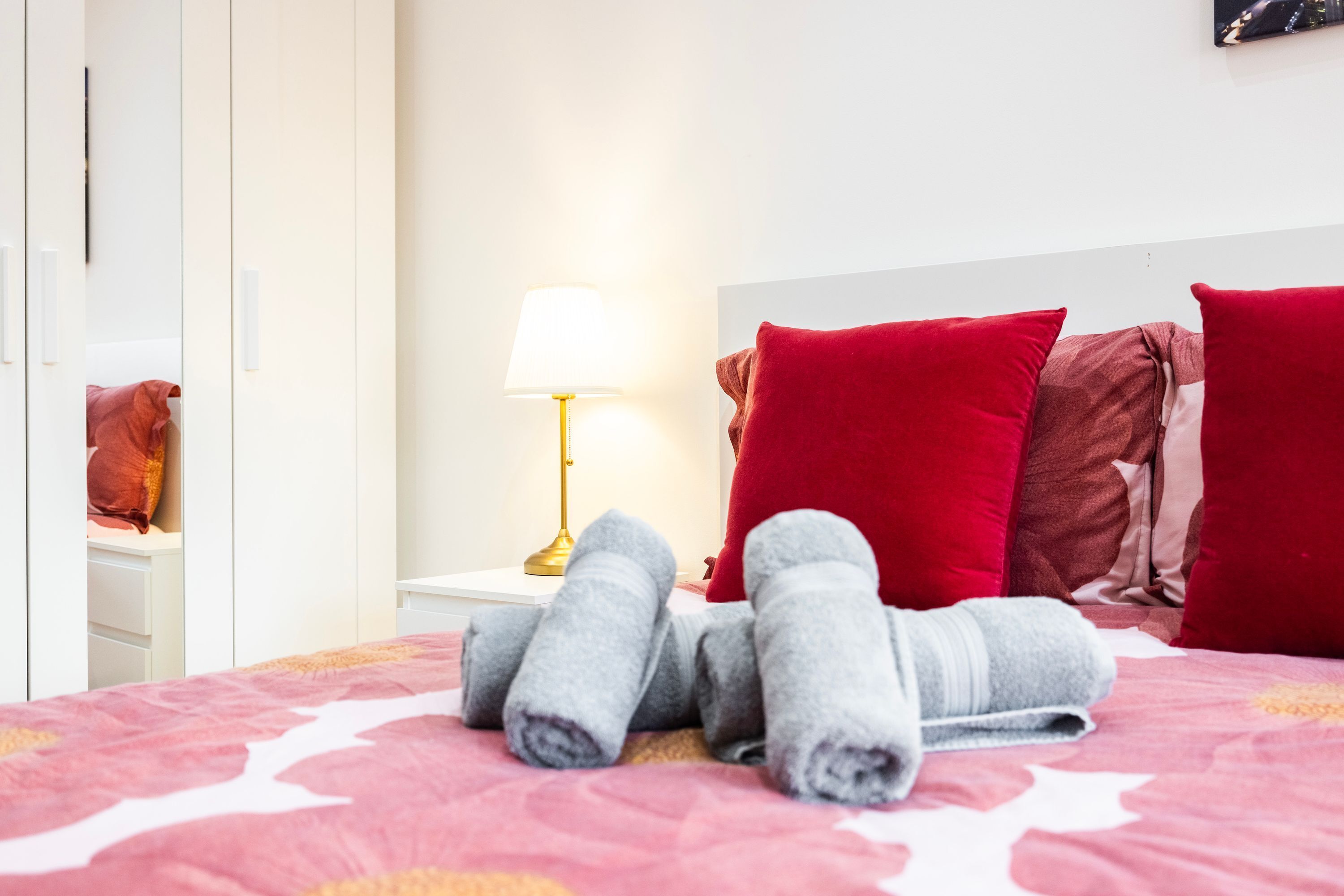 A neatly made bed with rolled towels, a lamp, and red pillows on a pink bedspread.