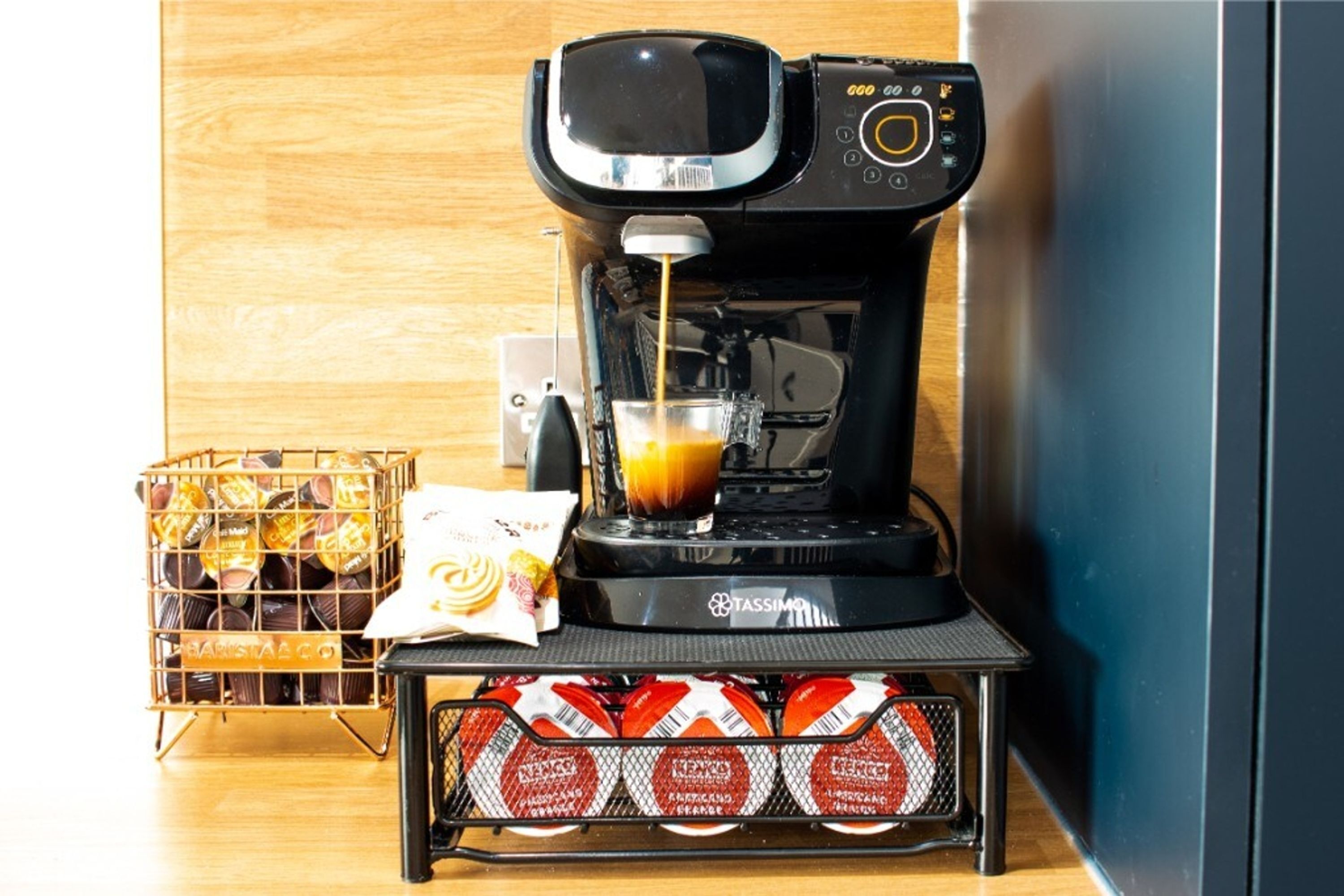 A coffee machine with a glass of coffee, a basket of pastries, and a container of coffee pods.