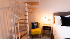 Moderni hotellihuone, jossa on kierreportaat, keltainen nojatuoli, yöpöytä ja valko- ja mustavärinen vuodevaatteilla varustettu sänky.