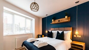 Moderni hotellihuone, jossa on suuri sänky, yöpöydät, ikkuna, jossa on sälekaihtimet, seinälle kiinnitetty hylly, jossa on kasveja, ja riippuvalaisin.