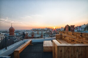 Une terrasse sur le toit, avec des bancs en bois et une balustrade donnant sur un panorama urbain au coucher du soleil.