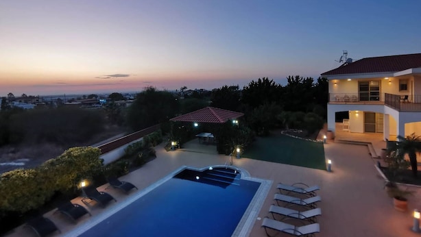 Une grande piscine avec des chaises longues, un patio couvert et une vue sur le paysage urbain au crépuscule.