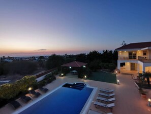 A large swimming pool with lounge chairs, a covered patio, and a view of a cityscape at dusk. - Orchard Blossom Private Villa With Pool (Frenaros)