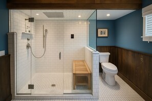 Een badkamer met een douchecabine van glas, witte betegelde muren, een houten bankje, een toilet en een ingelijst foto aan de muur.