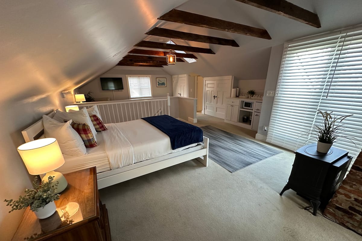 Ein Schlafzimmer mit einem Bett, Nachttisch, Lampe und einem kleinen Topfpflanzen. Ein Fenster mit Jalousien bietet einen Blick nach draußen.