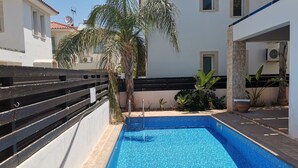 A swimming pool with tiled blue floor, surrounded by a wooden fence, palm trees, and a building with a tiled roof. - Amelia Villa With Private Pool Wifi BBQ (Paralimni)