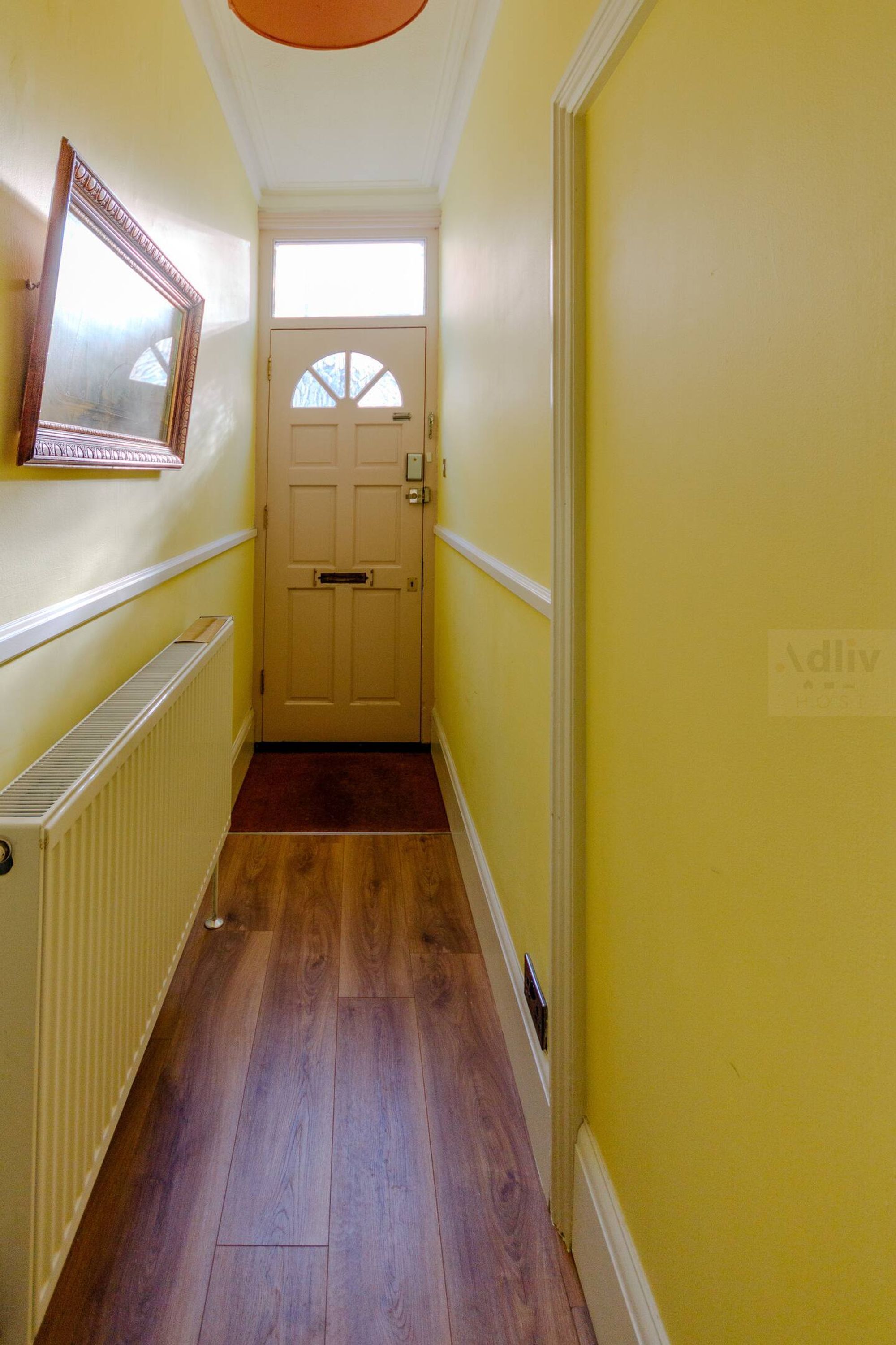 A narrow hallway with yellow walls, a wooden floor, a white door with a window, and a radiator on the left.