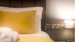 Ein ordentlich bezogenes Bett mit einem gelben Kopfkissen und einer weißen Decke, ein Nachttisch mit einer Lampe und eine Steckdose.