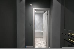 Un couloir étroit avec une porte donnant sur une salle de bain dont le mur derrière le lavabo est recouvert de carreaux blancs disposés en motif chevron, et dont le sol présente un motif géométrique.