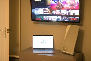 A television displaying a movie streaming service interface with a laptop and a white device on a table.