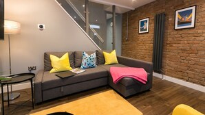 Ein modernes Wohnzimmer mit grauem Sofa, gelb- und mehrfarbigen Kissen, einer rosafarbenen Decke, einer Glasstufenkonstruktion und einer Ziegelwand mit zwei gerahmten Bildern.