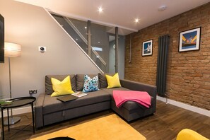 Un salon moderne avec un canapé gris, des coussins jaunes et multicolores, un plaid rose, un escalier en verre et un mur de briques orné de deux tableaux.