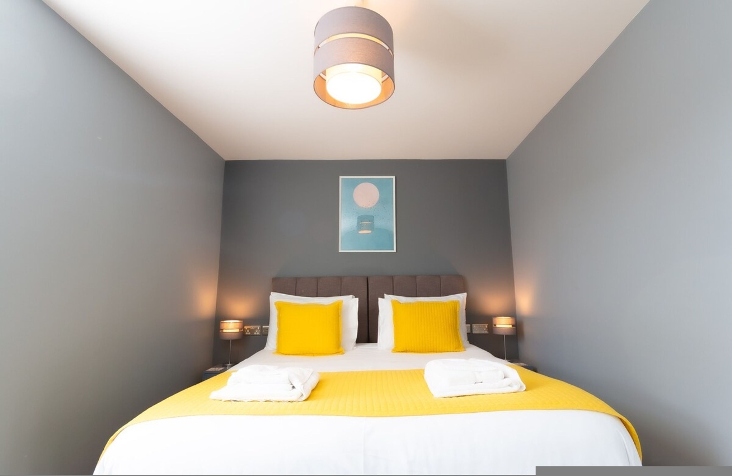 Quarto de hotel com uma cama, duas luminárias laterais, uma luz no teto e um quadro pendurado na parede.