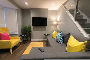 Un salon moderne avec un canapé gris, des coussins jaunes, un fauteuil jaune, un téléviseur à écran plat et un escalier.