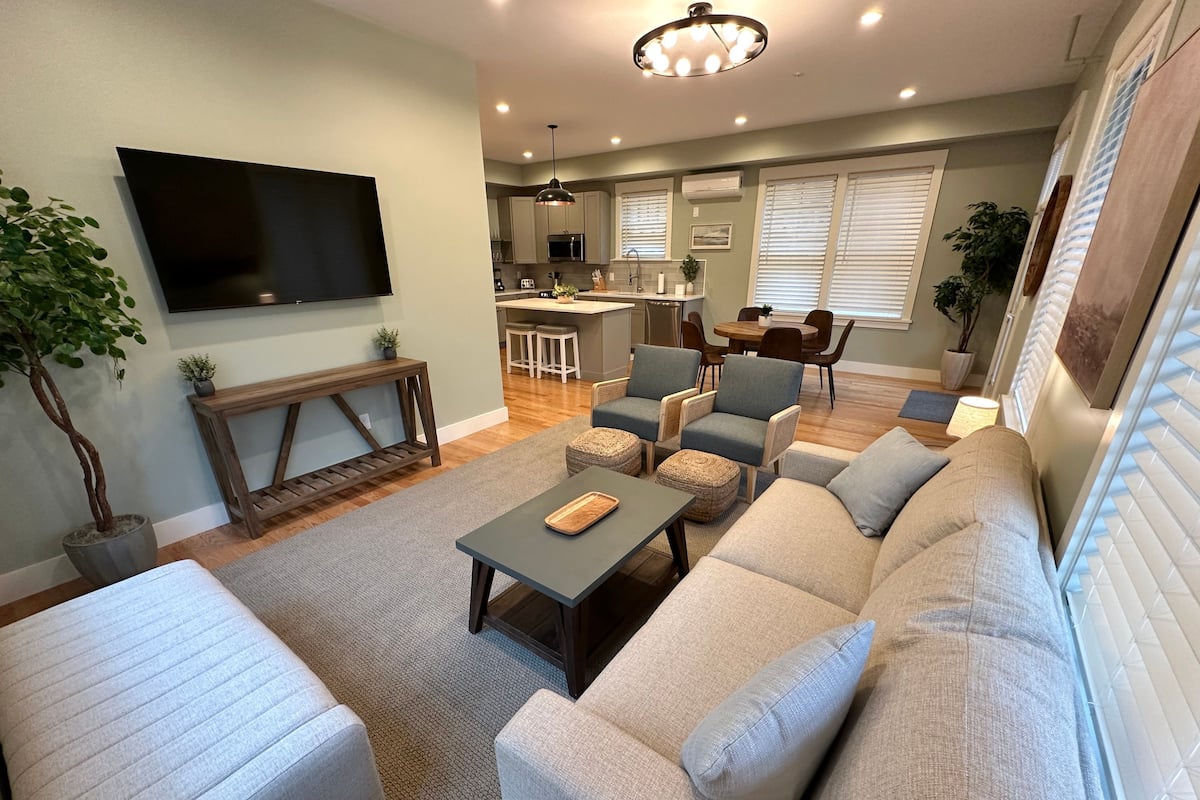 A modern living room with a sectional sofa, a coffee table, and a TV mounted on the wall. There is a dining area with a table and chairs, and a kitchen in the background.