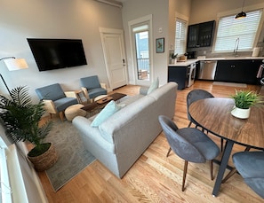 A modern living room with a grey sofa, blue armchairs, a wooden coffee table, and a dining area with a round table and chairs. - Brand New Construction! 3 Unit/5 Bed Property (Newport)