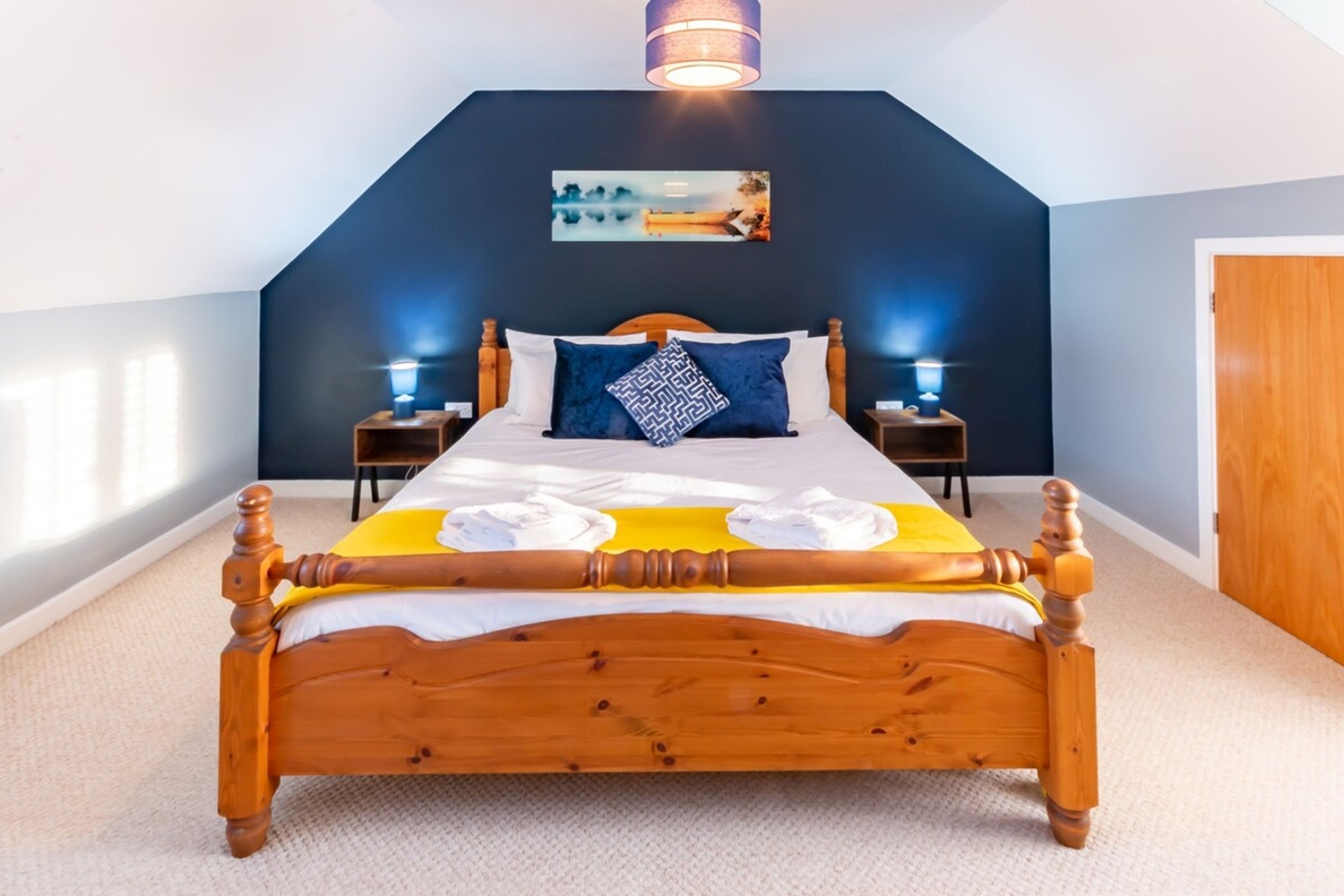 Quarto com cama de madeira, duas mesas de cabeceira, um criado-mudo, uma luminária na parede e uma porta.