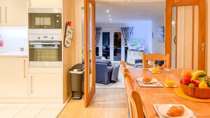A modern kitchen with built-in appliances, a dining area with a wooden table set for breakfast, and a view into the living room. - Stylish Margate Coastal Home Parking By Adliv (Margate)