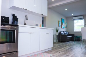 A modern kitchen with white cabinets, a stainless steel oven, and a sink. There is a black sofa and a glass coffee table in the living area. - Flamingo Beach House (Dania Beach)